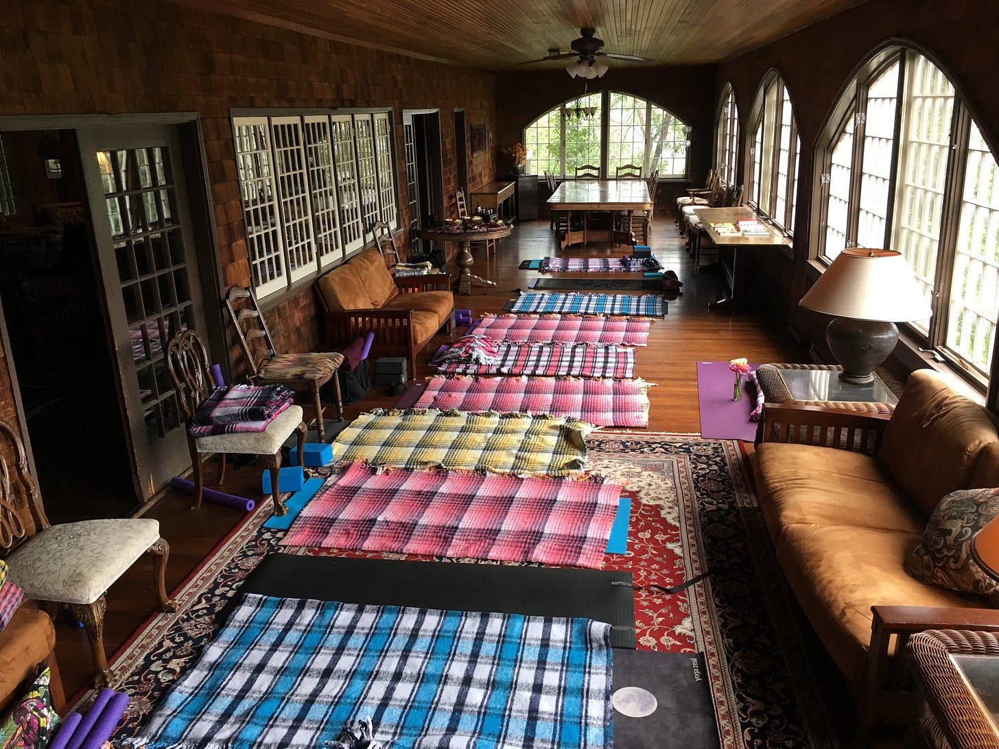 A room inside a wooden cabin, with yoga mats and blankets spread out next to each other on the floor. Chairs, couches, tables, and lamps fill the room, as well.