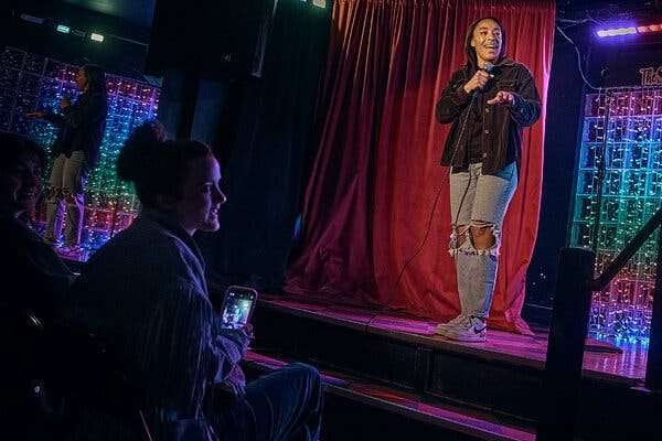 A woman wearing jeans and a button up jacket stands on a stage and smiles while holding a microphone. She gestures with one hand as audience members look on. A red curtain is in the background. 