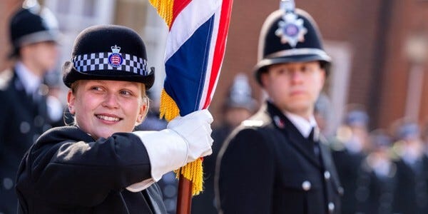 Female officer in ceremonial uniform holding colours while other police colleagues pass out to become police officers.