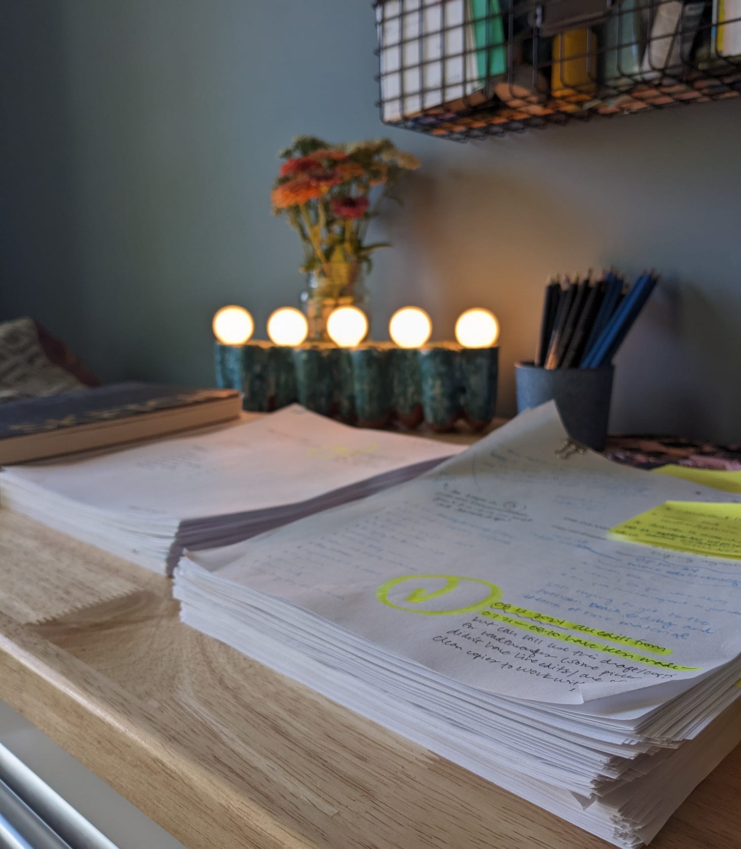 Three piles of paper and notebooks on my wooden desk, with a small lamp in the background with five lights, against a blue wall.