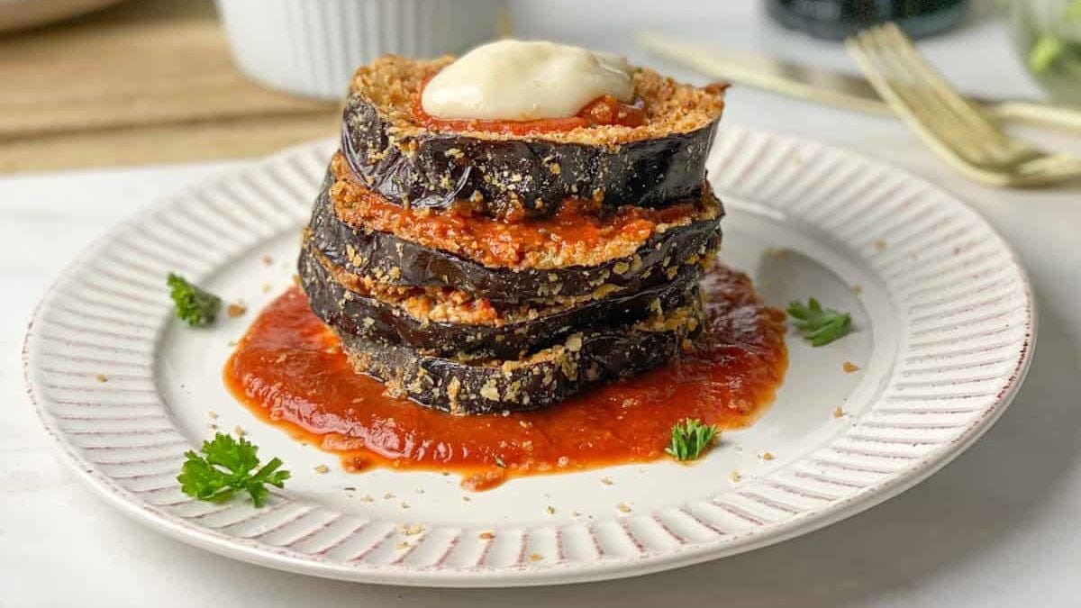 A stack of eggplant slices layered with tomato sauce on a white plate, topped with a dollop of cream, garnished with parsley, on a wooden table.
