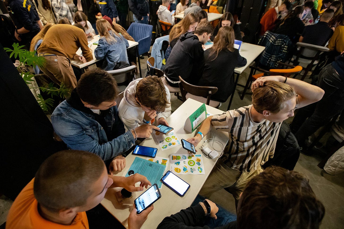 Groups of students sit at tables while holding mobile phones and examining paper documents and fliers.