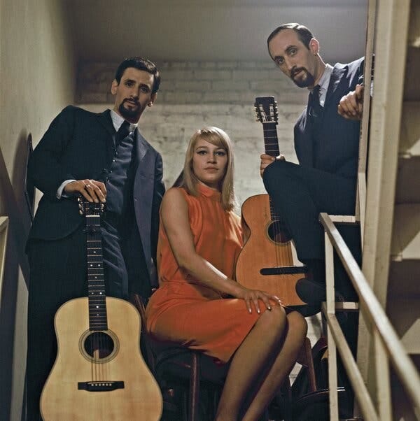 Two young men holding acoustic guitars stand on either side of a young woman, who is sitting on what looks like a suitcase or a trunk. They are on a stairway in front of a brick wall.