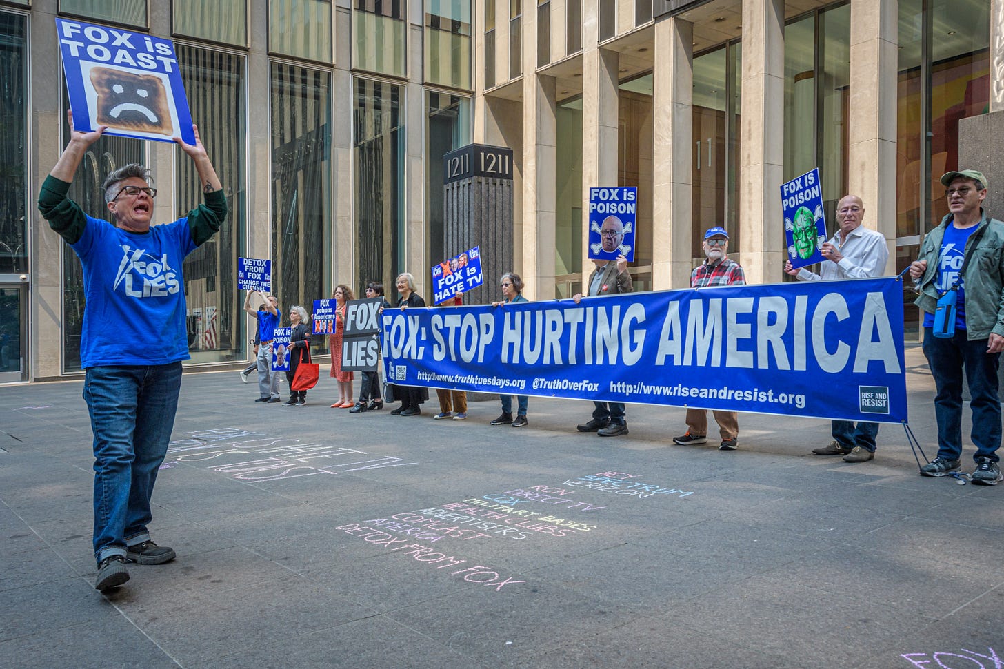 A dozen protestors, several holding a banner that reads  "Fox: Stop Hurting America" stand outside a concrete and glass building. In the foreground one protestor holds a sign reading "Fox is Toast"