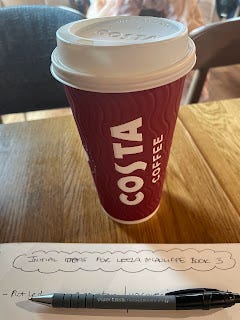 A view of a costa take away cup on a table. In front of the cup is a notepad and biro. The title of the page is just visible. It says 'Initial Ideas for Leeza McAuliffe Book 3.'