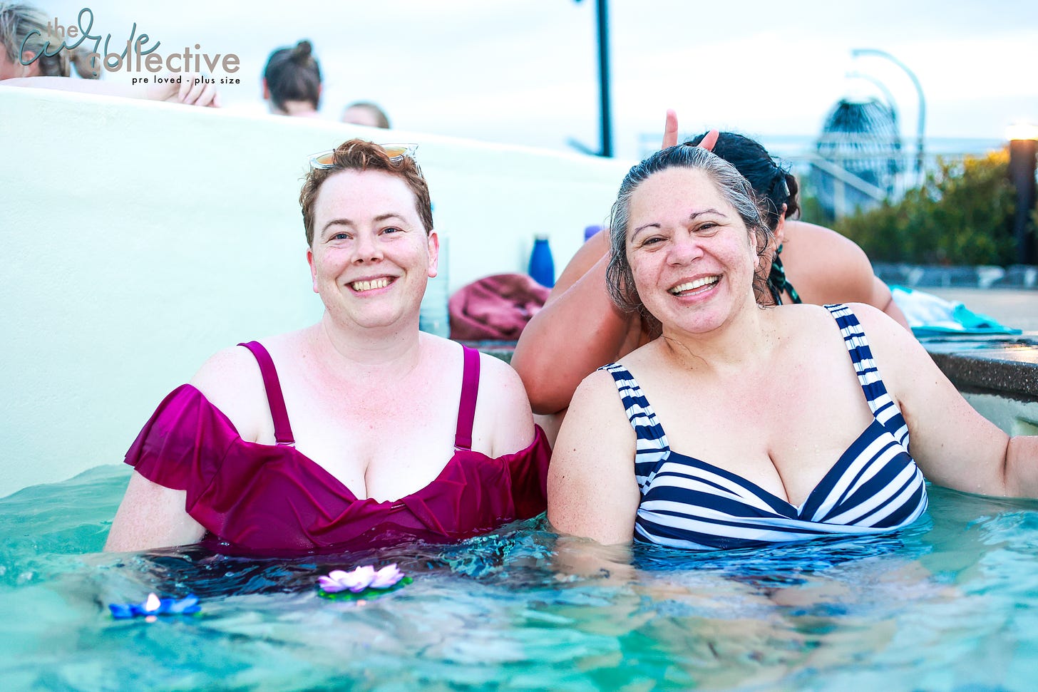 A fat Pakeha woman in a raspberry pink swimsuit with absurd and delightful ruffles, and a Māori woman in a navy and white striped swimsuit (so chic, super French) stand cleavage deep in water, clearly having a terrific time. Behind the white-and-navy suited woman, a MYSTERIOUS STRANGER has hidden her face, but holds up her fingers in bunny ears.
