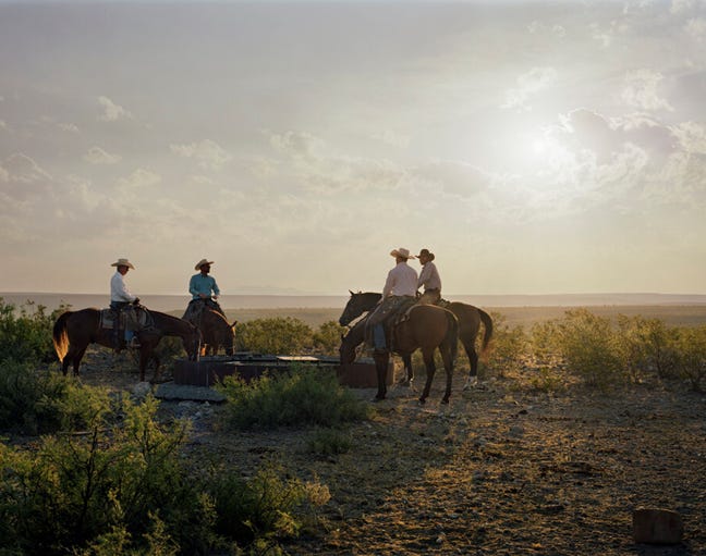 Image 2: color photo, huge sky, flat desert range land of Texas, four cowboys mounted on four horses pause at a watering trough. The cowboys are all Mexican nationals who legally work in Texas. 