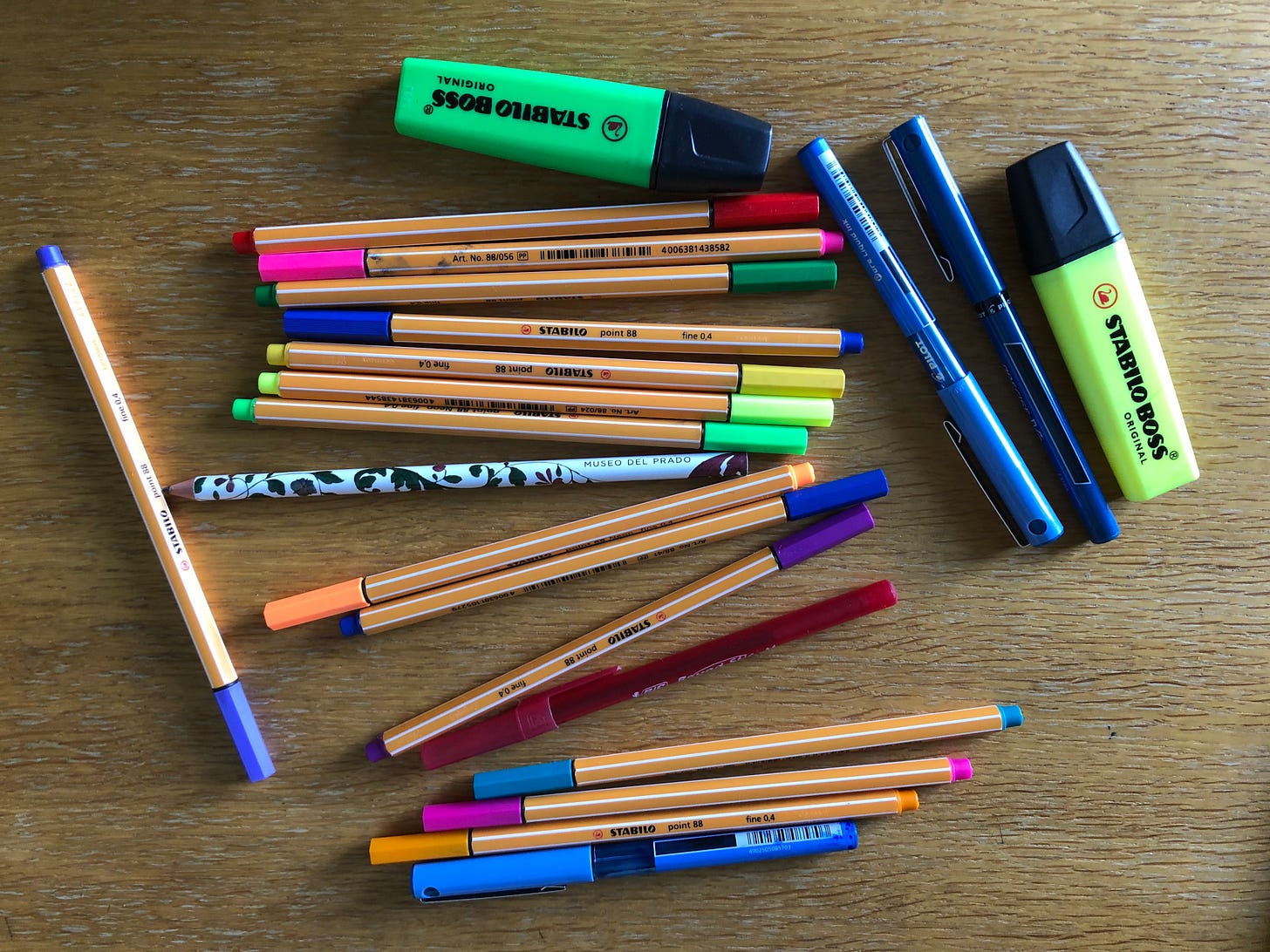 An array of pens, highlighters, and a pencil are jumbled on a wooden table.