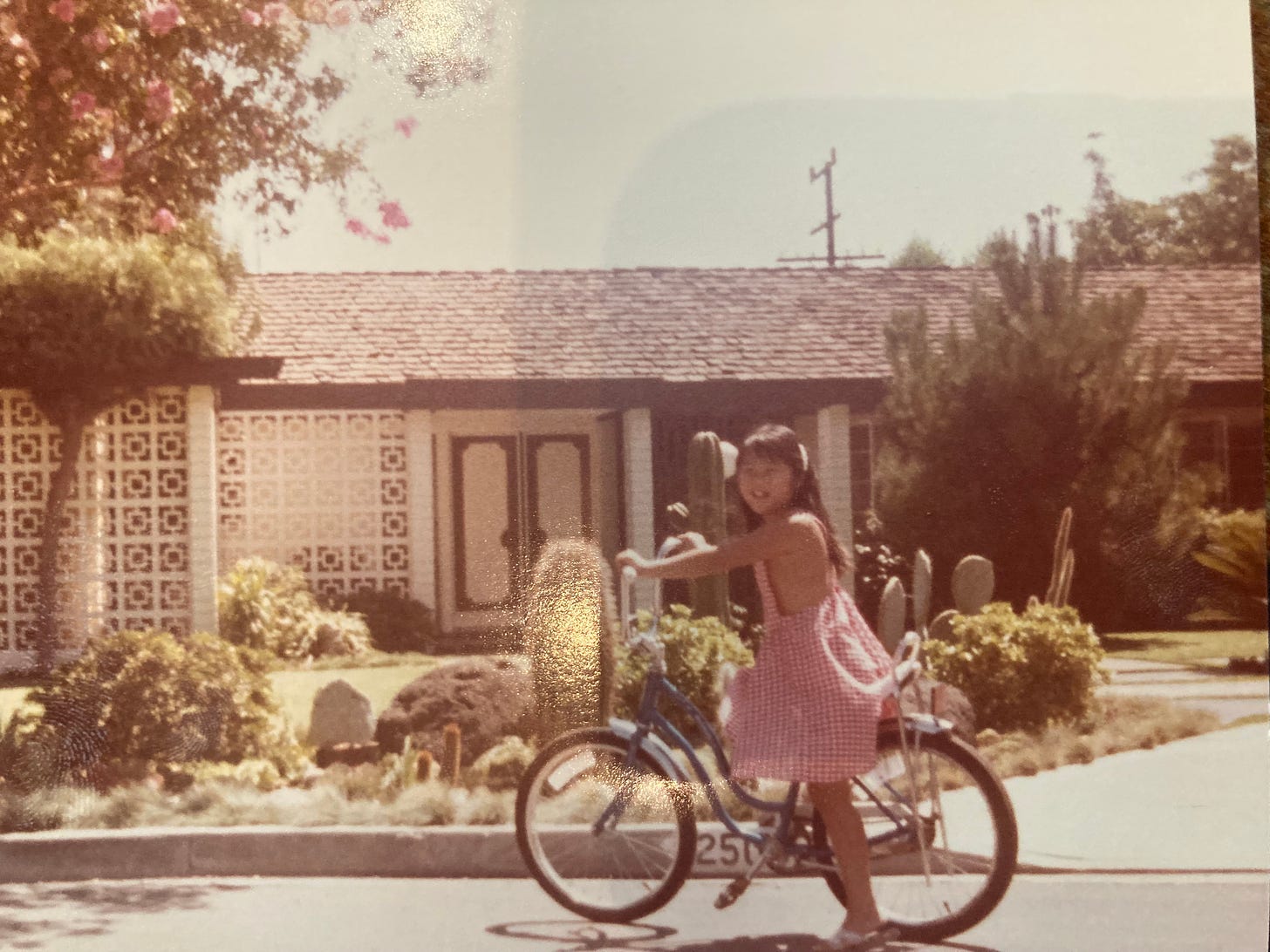 picture of me, as a school aged girl, in a red gingham pinafore and a bicycle in front of my childhood house with facade and original doors and landscaping.