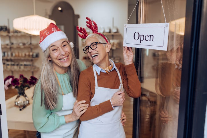 Two female business owners stand in their shop front wearing festive antlers and santa hat. They're laughing and holding the shops "open" sign.