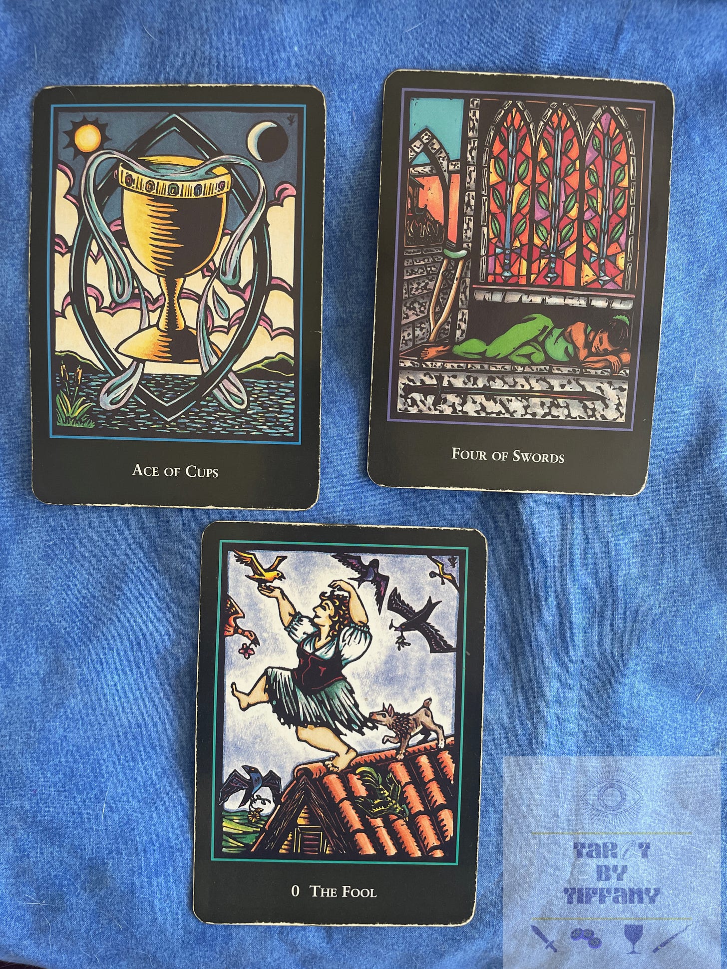 3-card spread using the World Spirit Tarot by Lauren O'Leary. The three cards are against a blue cloth background. Top two cards (left to right): Ace of Cups and 4 of Swords. Bottom card: The Fool.