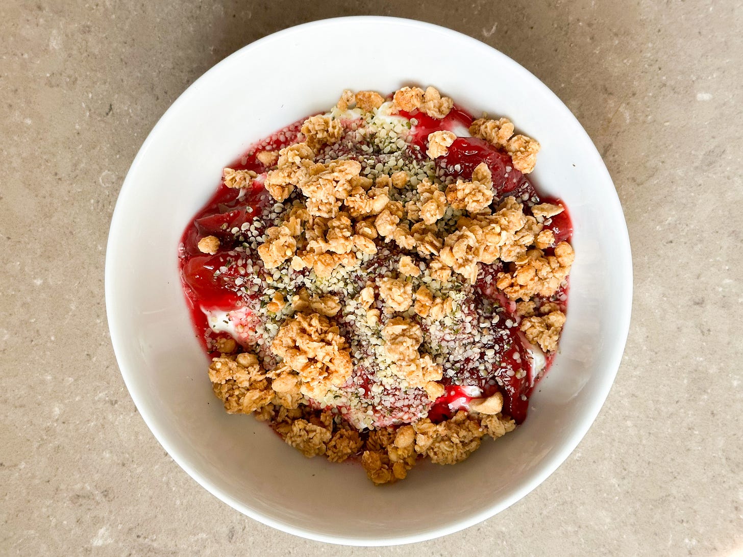 A photo of a white bowl with Greek yogurt, roasted plums, hemp hearts, and granola.