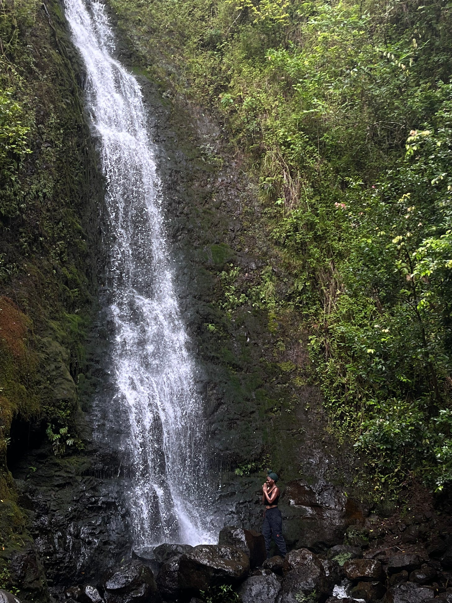 A photo of a woman smiling toward a steep waterfall in Hawaii with palms faced together