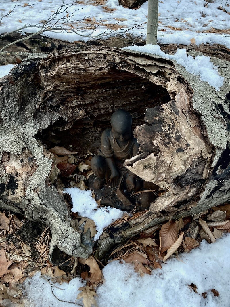 a snowy forest floor with a hollowed-out log. a small buddha statue is seated in the shadows of the log.