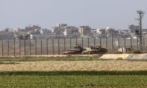 Israeli troops and tanks are stationed in front of a high wire fence across a field; a ruined city is in the background