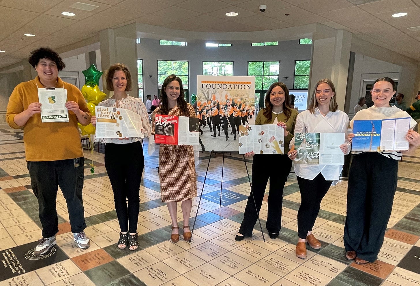 Six smiling people hold magazines in a light-filled room.