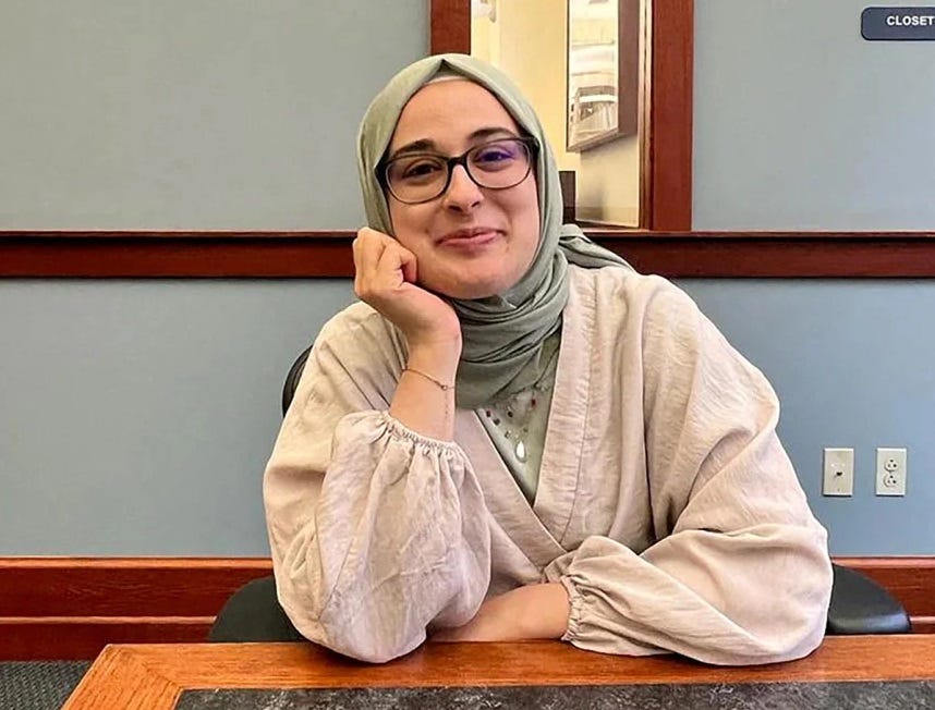 Rümeysa Öztürk sits at a table in an unidentified building (Courthouse? University seminar room?), wearing glasses, a light tan long-sleeved outfit and light-green hijab. She smiles slightly, one hand cupped against her chin.