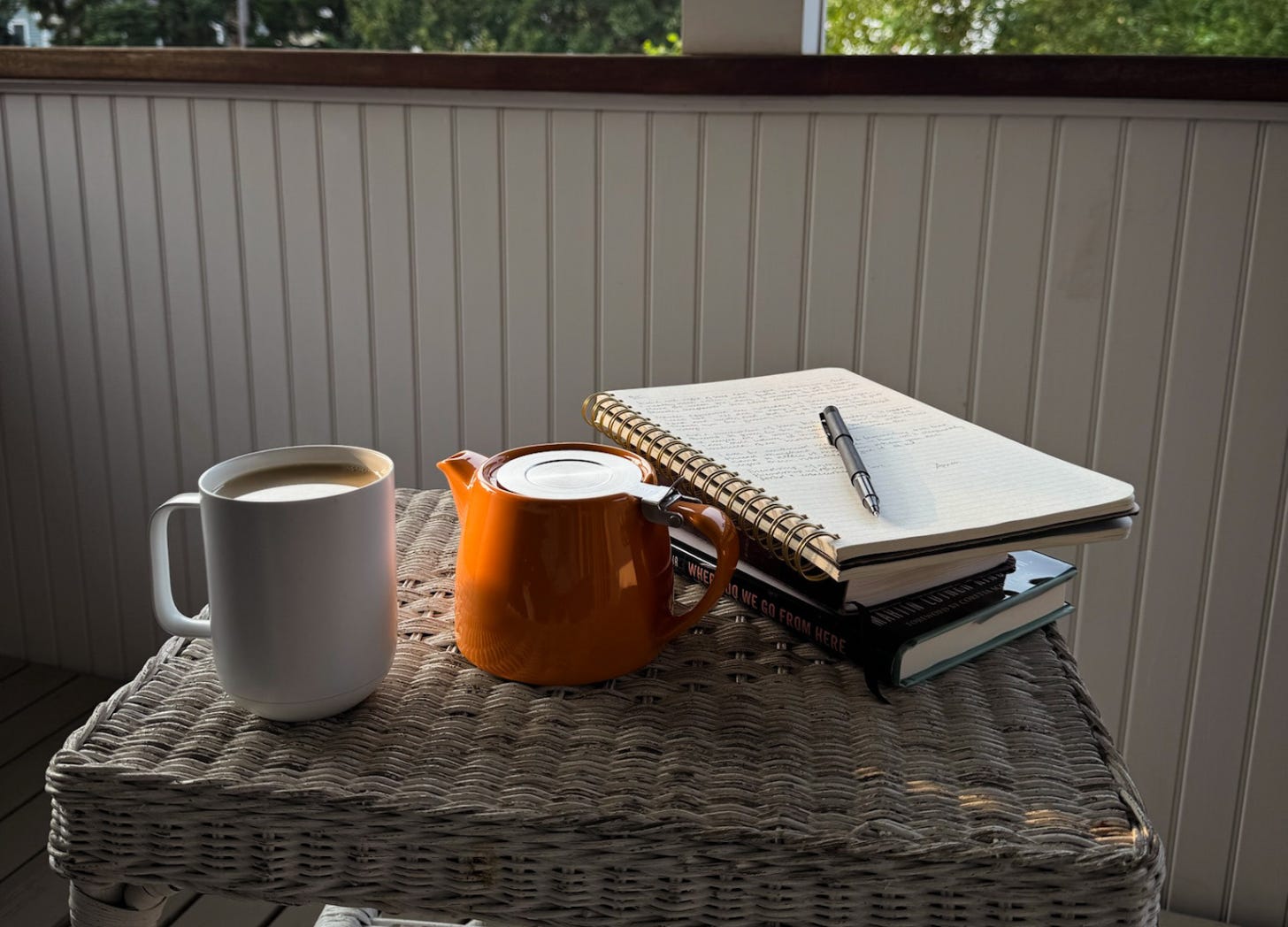 photo of a full mug, tea pot, and stack of notebooks and books on a wicker table outside on a porch