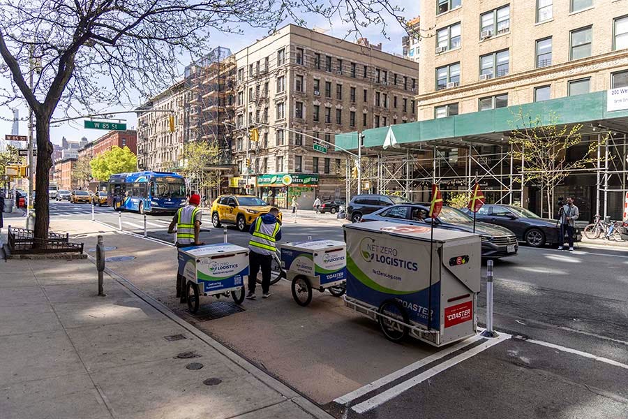 Two delivery workers place packages in cargo bikes parked in a curb extension along a busy roadway on the Upper West Side of Manhattan.