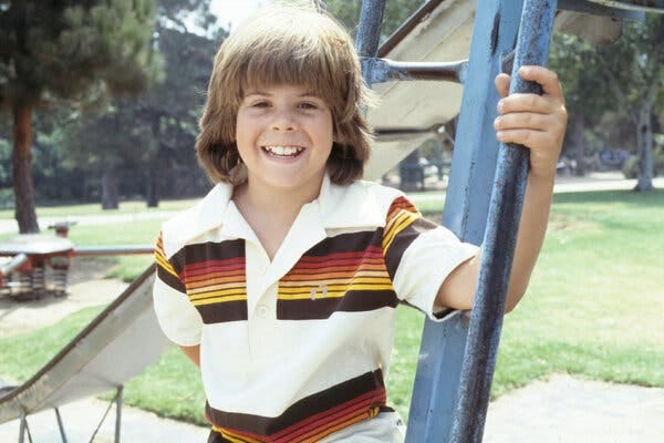 Adam Rich, wearing a polo shirt, holds onto the metal pole of a playground slide.