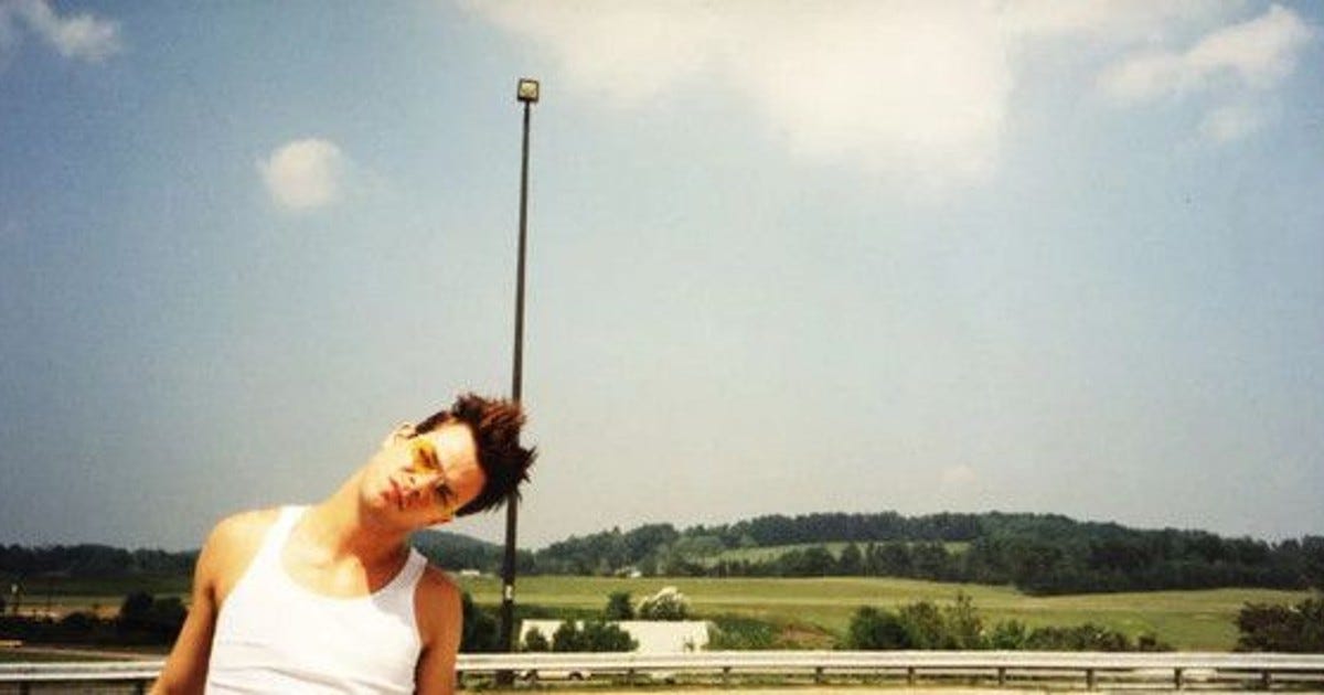 Movie still from Tarnation. A person in a tank top tilts their head to the side, standing outside against a field.