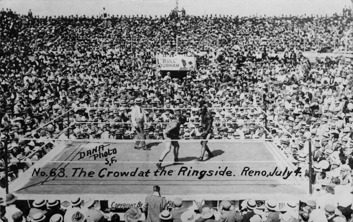 An aerial view of a crowded boxing ring with thousands of spectators wearing straw boater hats, captioned ‘The Crowd at the Ringside. Reno, July 4.’