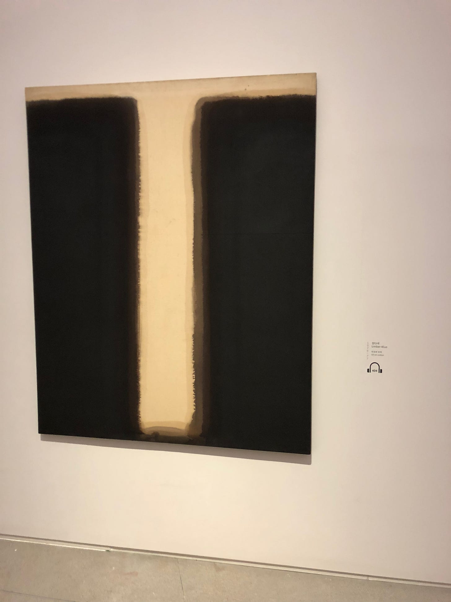 Umber-Blue by Yun Hyong-keun. The painting is oil on cotton. Two simple black bars tower over a narrow path, looking like a gate. The edges of the bar blur, making the gate seem like it’s just now opening, or closing. The path sprawled out before you is inviting but also holds a quiet menace of uncertainty.