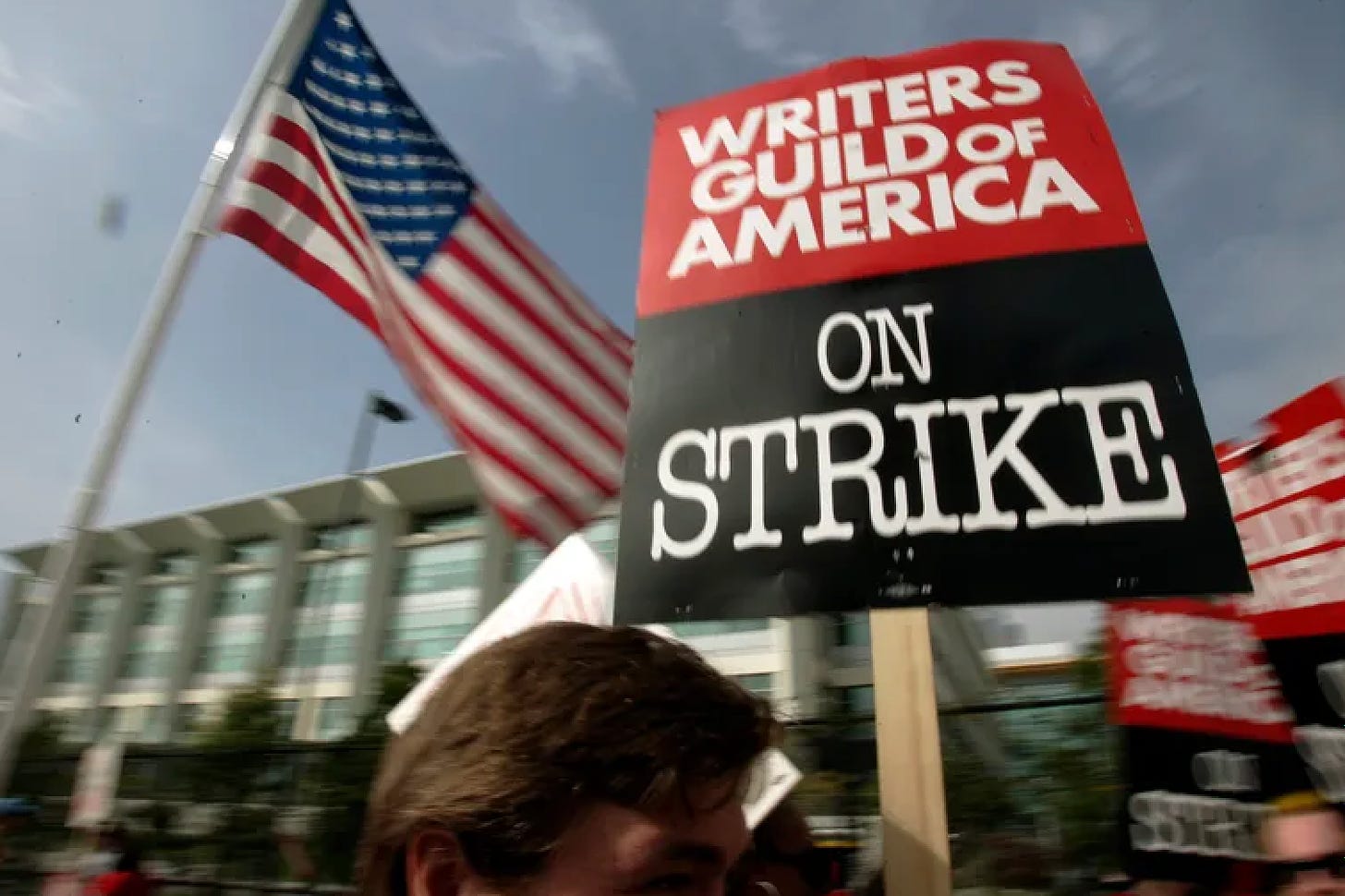 An image taken from a low angle of the WGA picket line. An American flag is waving in the background. The focal point is a sign being held in the air that reads "Writers Guild of America ON STRIKE." 