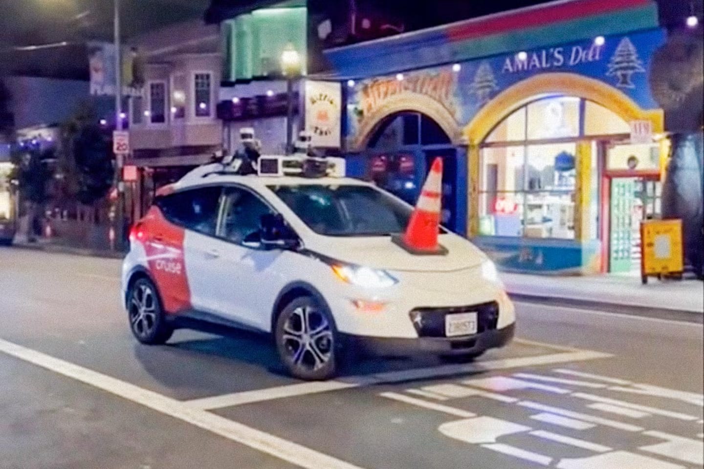 A white car stopped in the middle of a street. There's an orange traffic cone on its hood.