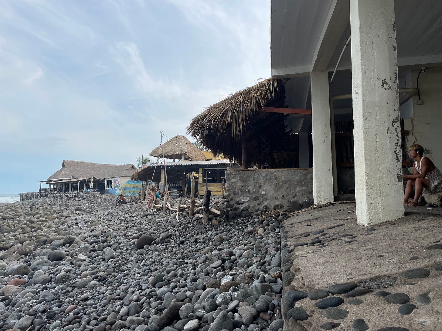 A man with headphone sits in a gutted building watching the ocean, with small thatch-roofed buildings beyond