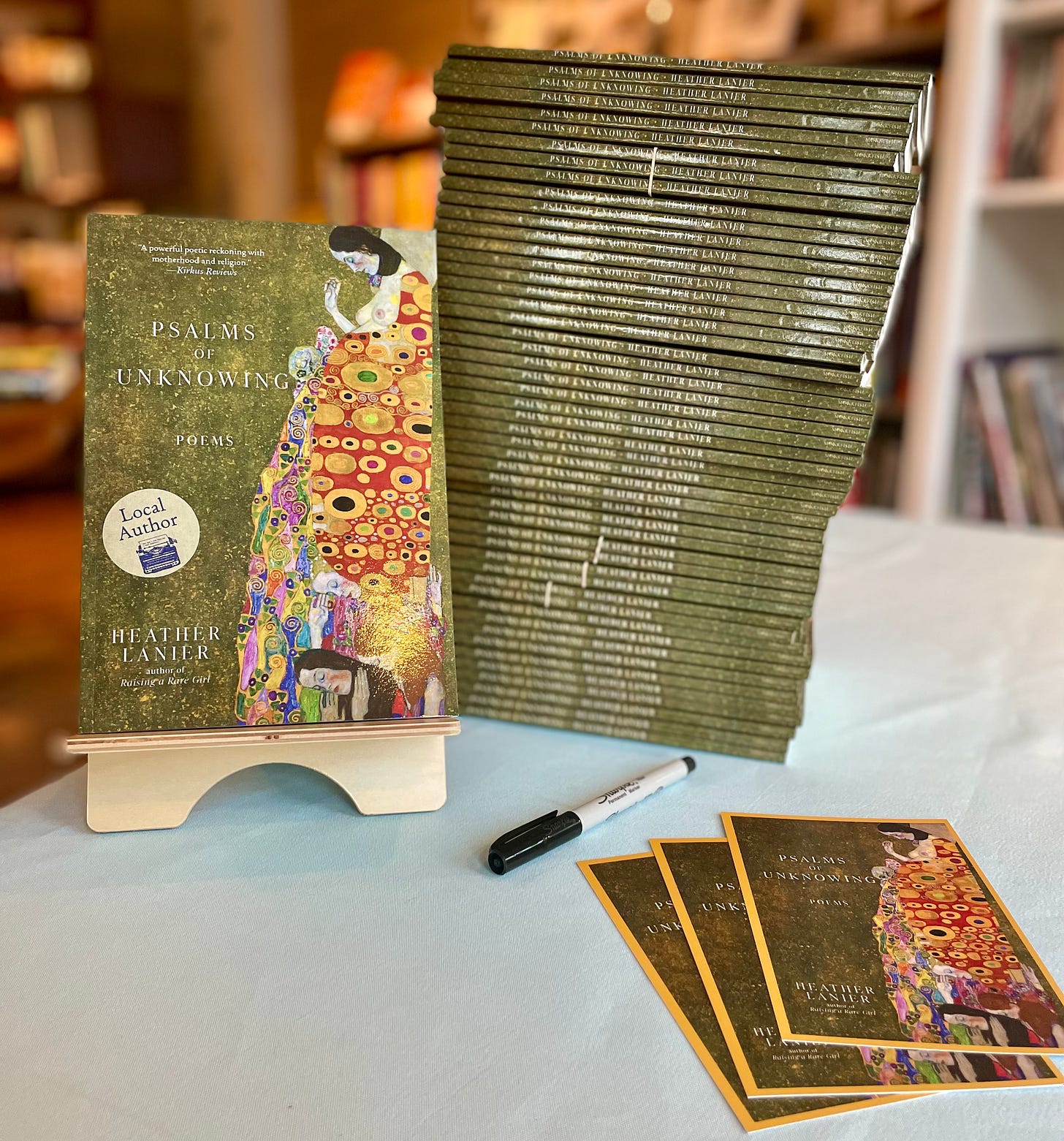 Table featuring a copy of Psalms of Unknowing, which features Klimt's HOPE II. Beside the book is a stack of 30 or so books with a green spine. In front of that stack are postcards with the same book cover displayed on the front, beside a pen.