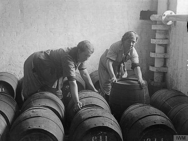 A black and white photo of two robust looking women shifting barrels. One is looking at the camera, as if inviting the camera operator to stop messing about and lend a hand.