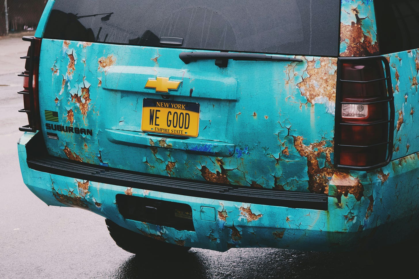 The back end of a rusted-out aqua-colored Chevrolet Suburban with a New York vanity license plate that reads "WE GOOD" 