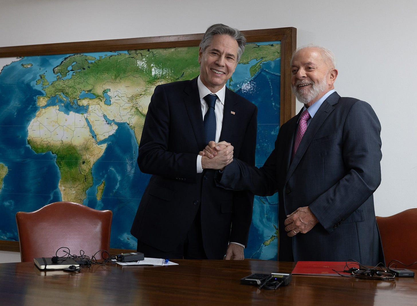 Secretary Blinken and Brazilian President Lula da Silva shake hands and smile for a photo while standing behind a wooden table.  A large world map hands on a white wall behind then.