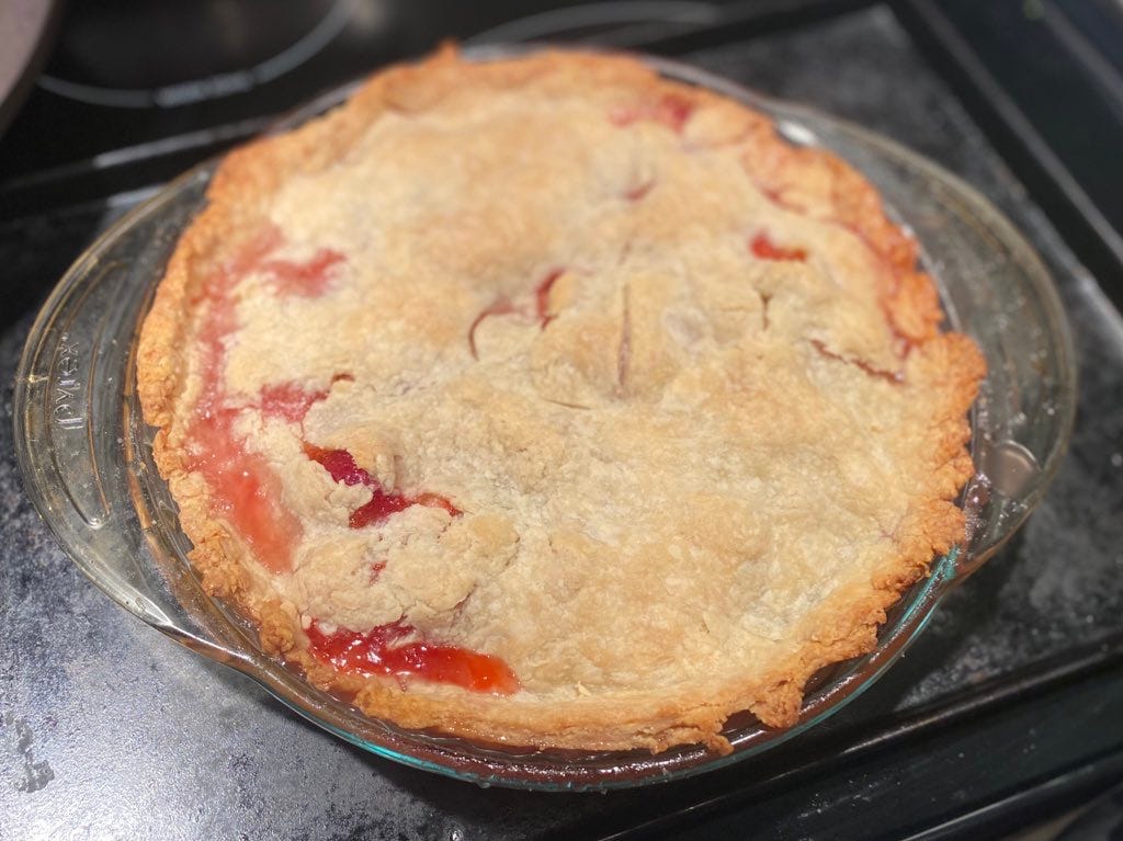 A pie with a browned crust and a heart cutout in the centre. The crust has cracked in several places around the edge, and pinkish-red juices have oozed over the top. The pie is in a clear glass pan on top of a baking sheet.