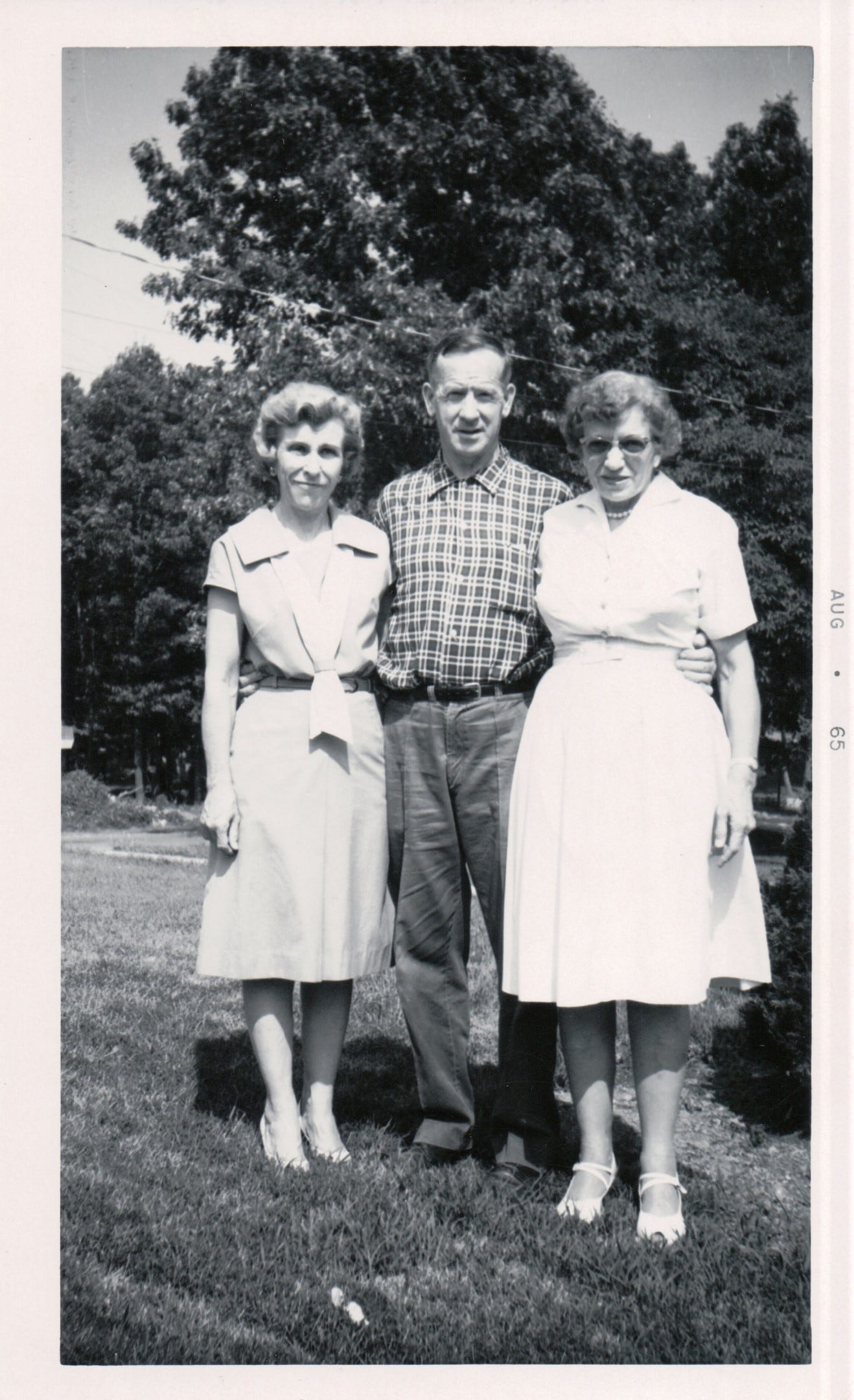 Two woman in casual dresses and dress shoes, and a man in a plaid shirt and dress pants posing for a photo outside