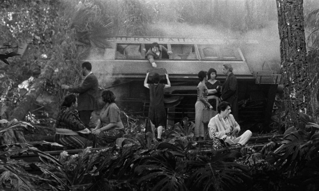 Movie still from Grand Tour. In the middle of a forest, people sit on the ground and help people with their luggage in a train