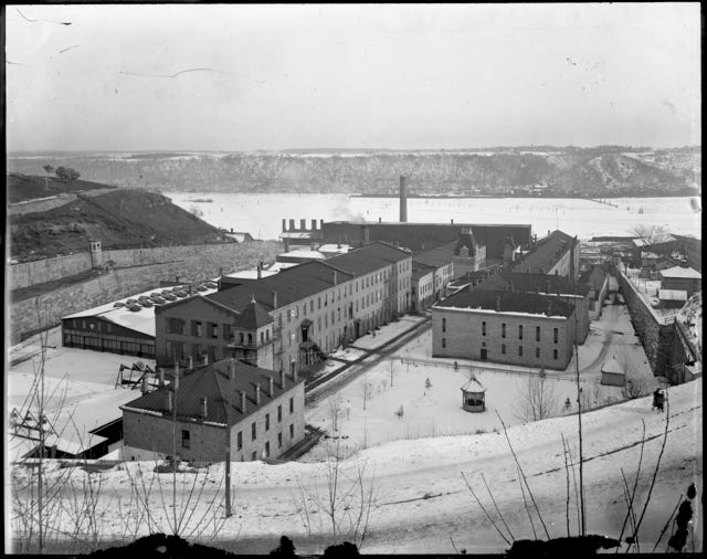 A greyscale archival photo with a black frame shows the view of a group of brick buildings on the shores of a frozen river during winter