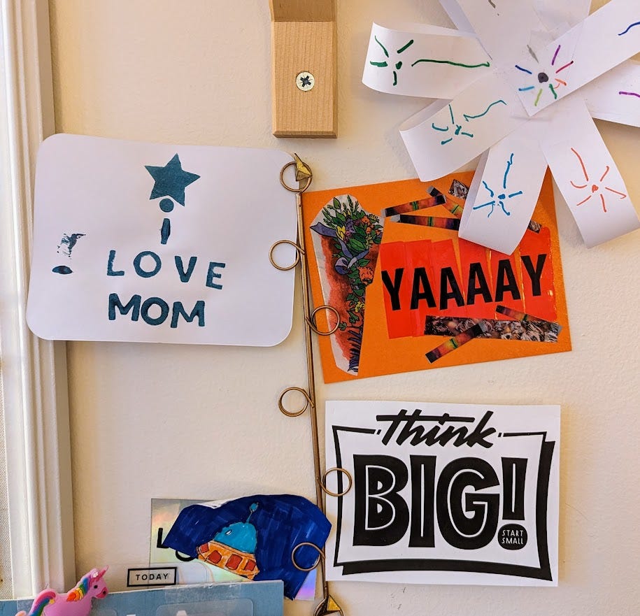art posted next to a writing desk--a postcard that reads I love mom, an orange postcard that reads YAAAAY, and a sign that says Think Big! start small