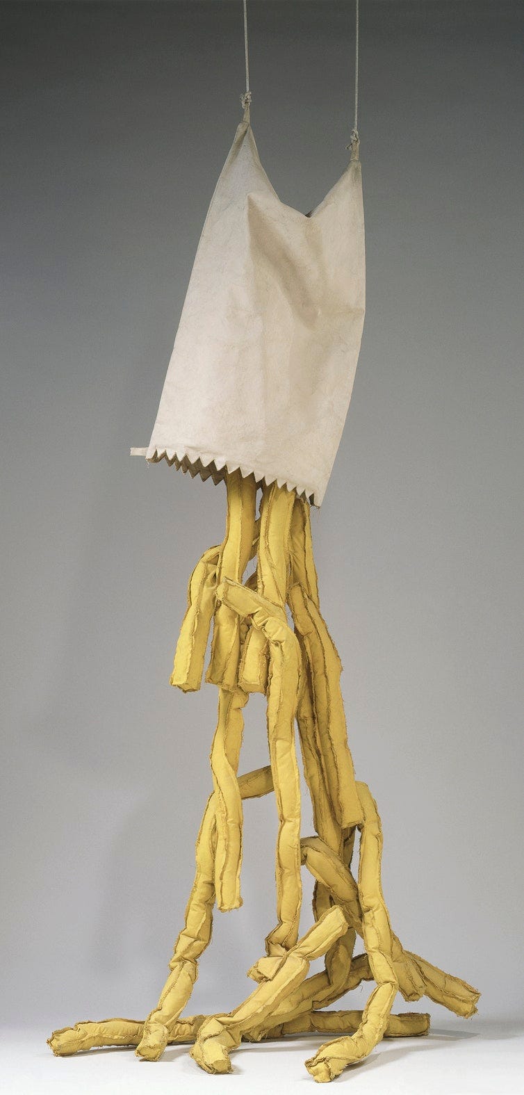 A rectangular cloth bag hangs with the mouth of the bag downwards, suspended by two cables. Long rectangular prisms ('fries') are falling from the bag's mouth. It looks like a drawing of a spilling paper bag of fries brought to life in massive scale.