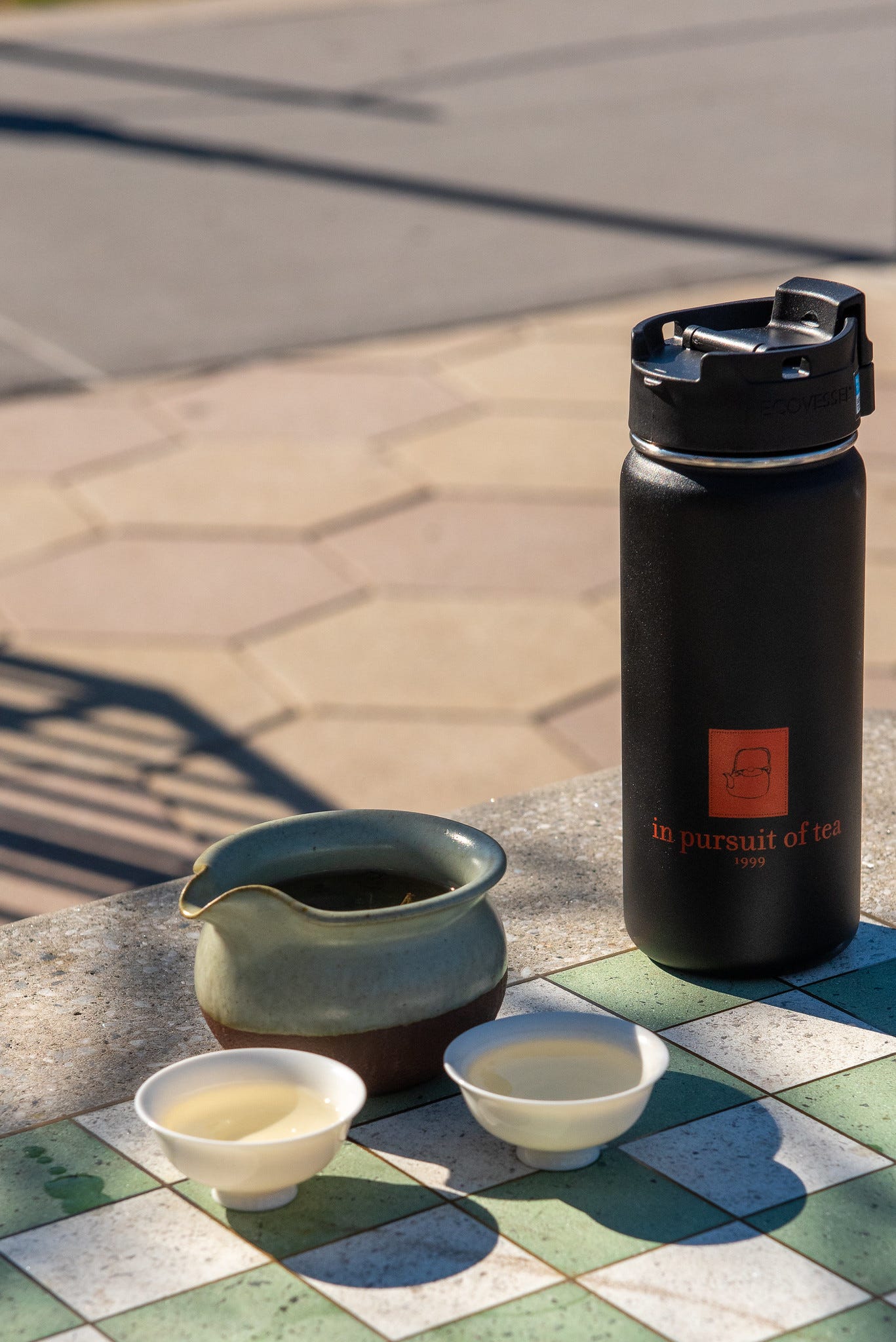 ID: Thermos and tea pitcher with 2 small cups of tea, out on a table in a park
