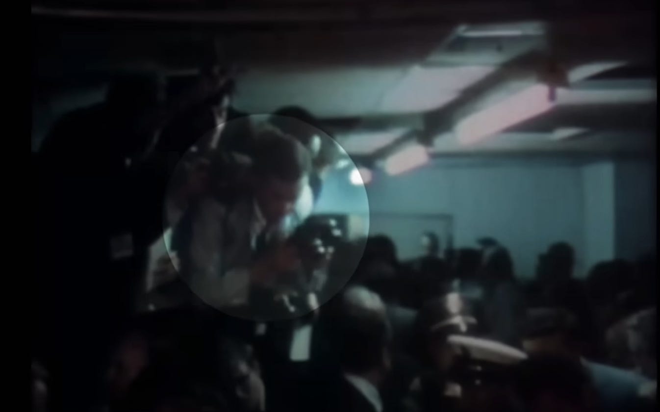 A blurry photo of a crowded kitchen hallway. A white man in a light suit is holding a camera. He is the focus on the picture.