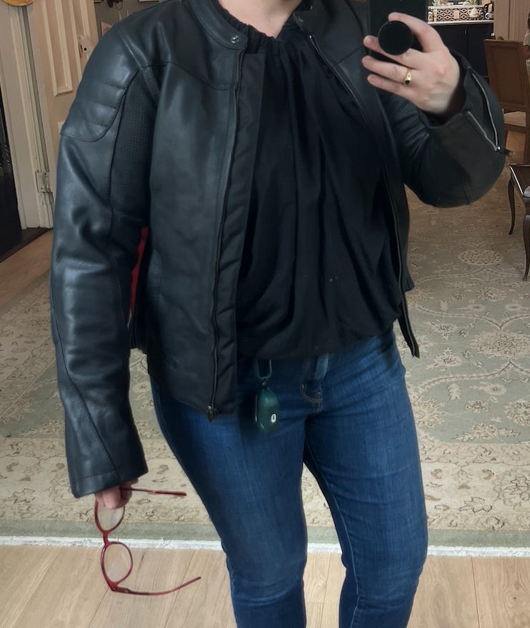 A mirror selfie of Amber from the neck down. She wears a black motorcycle jacket and blue jeans. She holds her red reading glasses in one hand, the other holds her cell phone which is taking the photo.