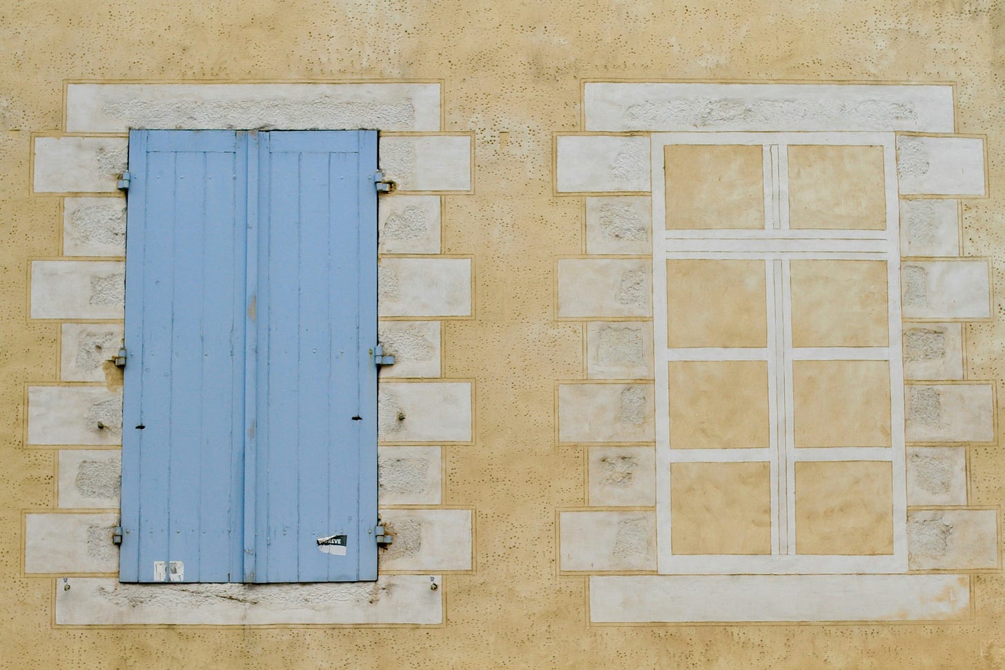 Parete esterna di un edificio di color giallino. Vi compaiono due finestre: a destra una finestra disegnata in bianco, con lo sfondo giallino del muro; a sinistra una finestra chiusa da imposte di legno color azzurrino.