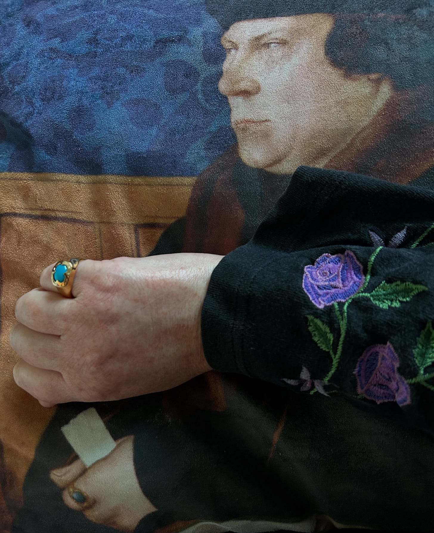 A hand wearing a gold ring containing a turquoise. The hand is resting on a cushion showing Thomas Cromwell, also wearing a turquoise ring.