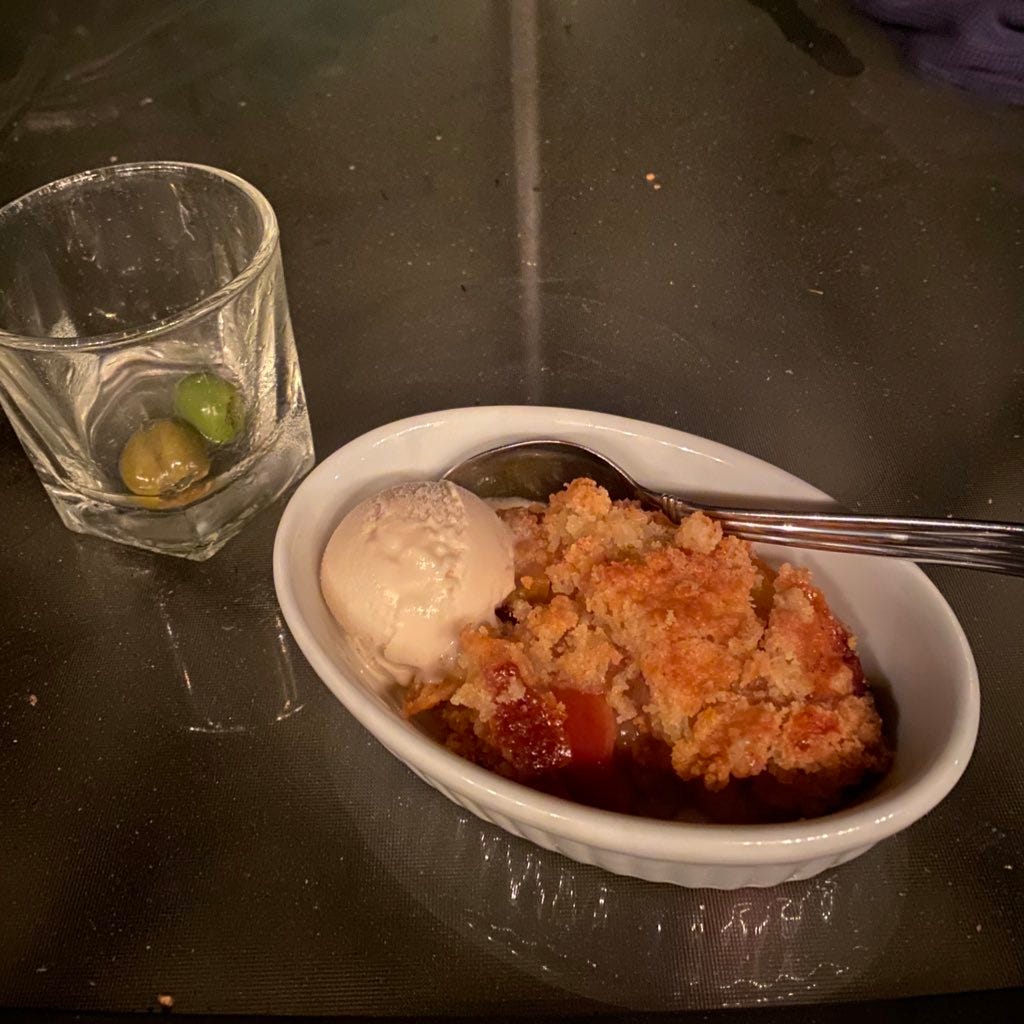A rocks glass with a nearly-empty martini, two kinds of olives in the bottom, next to a small oval dish of cobbler. A scoop of vanilla ice cream is at the left edge of the dish, and a spoon rests at the back. It's on an outdoor table, the photo clearly taken at night.