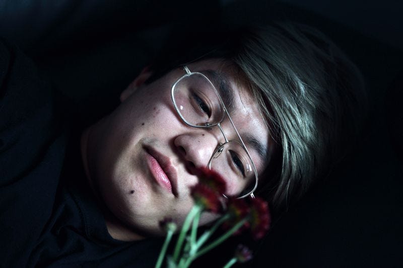 Person with green hair and glasses looking wistfully at the camera through a small bundle of flowers