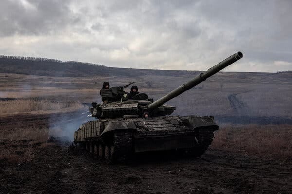 Soldiers man a tank that cuts a path through a brown, open field. Gray clouds hang in the sky.