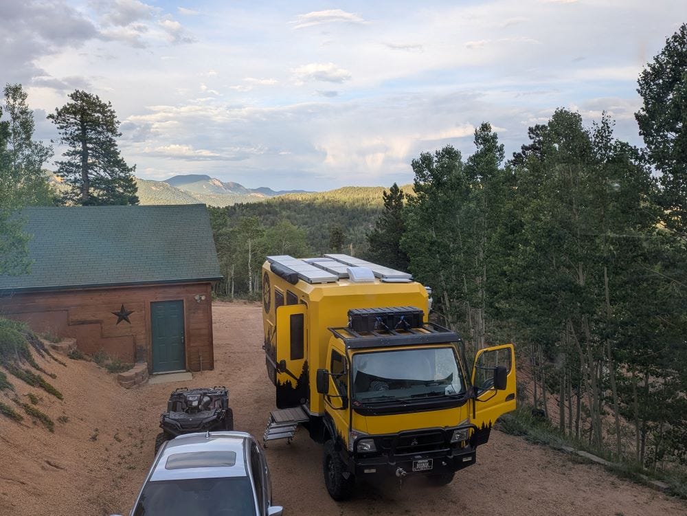 Walter and other vehicles in a dirt driveway with an outbuilding, and layers of mountains in the background