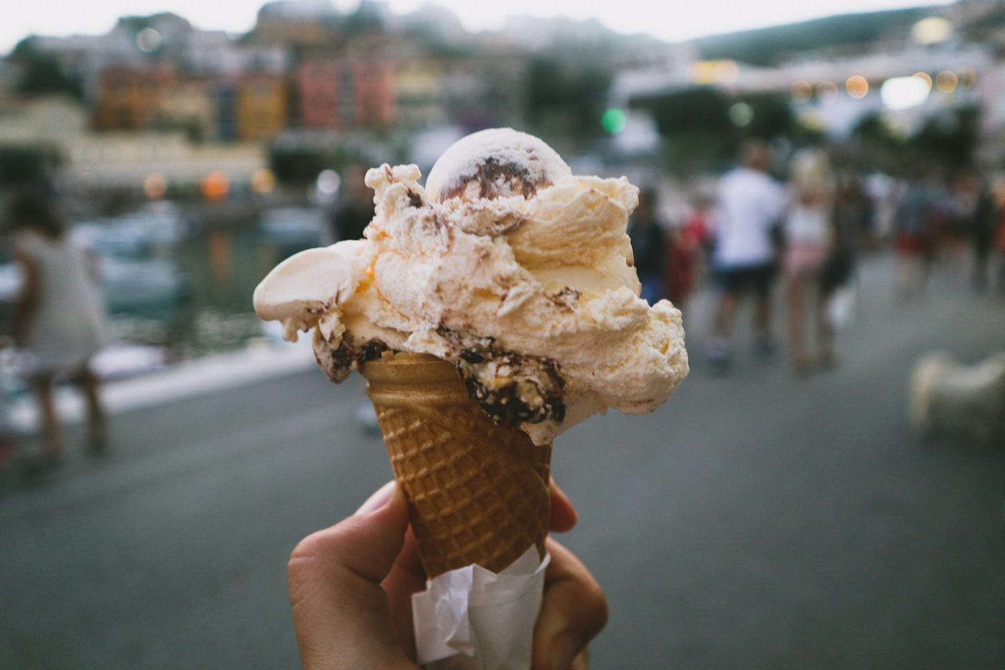 white ice cream with brown chocolate chunks in a waffle cone with a water front and pastel colored buildings in the background